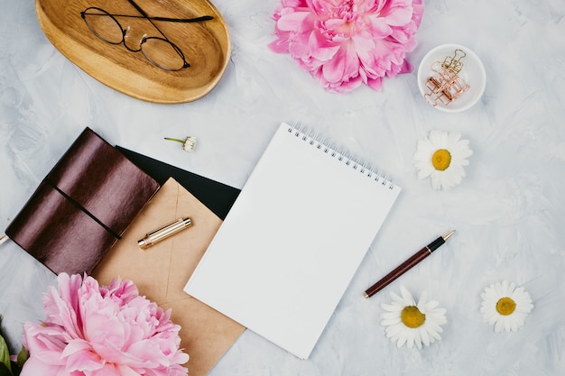 Maquete de negócios feminino com artigos de papelaria, margaridas, flores de peônia, cadernos e óculos, flatlay em fundo de cimento