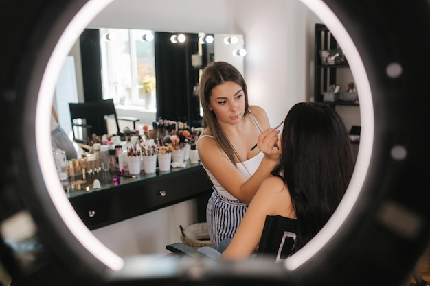 Maquiador fazendo uma maquiagem para uma jovem atraente Linda jovem morena Lâmpada de anel