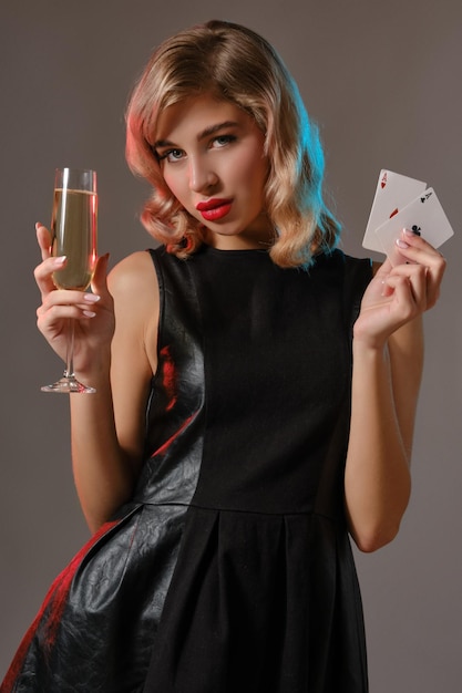 Maquiagem brilhante de mulher loira encaracolada elegante em vestido preto elegante segurando duas cartas de baralho e taça de champanhe posando contra o fundo cinza do estúdio Jogo de poker closeup