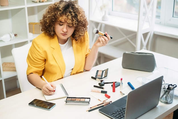 Maquiagem de escritório Trabalho multitarefa Asseio corporativo Vida de mulher de negócios Empregada ocupada com excesso de peso com cabelos cacheados na mesa aplicando cosméticos tomando notas no local de trabalho leve