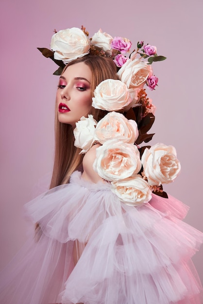 Maquiagem vermelha brilhante de mulher de beleza, flores rosas na cabeça de mulher. Art Girl em um vestido transparente de luz em um fundo rosa. Maquiagem brilhante profissional