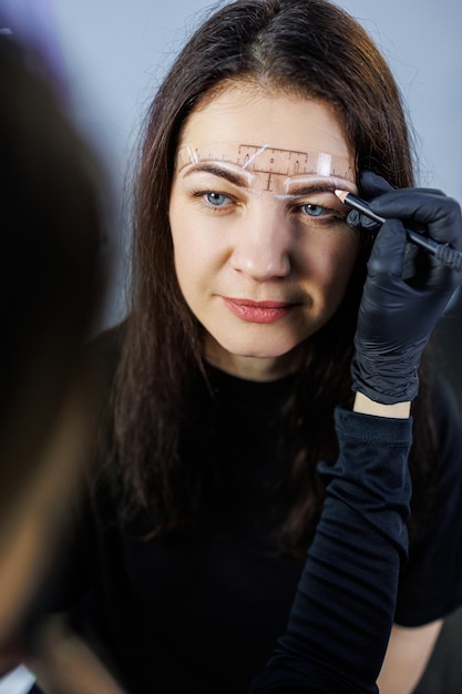 Un maquillador permanente marca las cejas de una mujer con un lápiz Procedimiento cometológico de maquillaje permanente Cuidado del rostro de una mujer