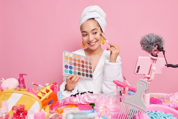 Maquilladora femenina muestra la transmisión en vivo de registros de paleta de sombras de ojos en línea a la audiencia desde casa rodeada de diferentes productos cosméticos y muestra cómo hacer el maquillaje diario. Blogger influyente