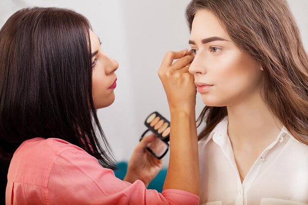 Maquilladora haciendo maquillaje profesional de mujer joven cerca del espejo en estudio de belleza