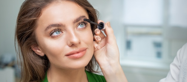 Maquilladora profesional aplicando rímel en las pestañas de la hermosa joven caucásica en salón de belleza