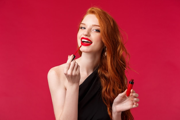 Maquillaje, belleza y concepto de mujer. Retrato de primer plano mujer pelirroja hermosa mujer aplicar lápiz labial, brillo labial rojo y mirando como en el espejo, de pie sobre una pared roja