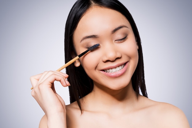 Maquillaje de belleza. Hermosa mujer asiática joven y sin camisa sosteniendo un pincel de maquillaje y sonriendo mientras está de pie contra el fondo gris