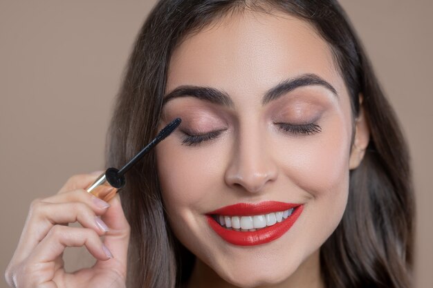 Maquillaje de día. Feliz sonriente hermosa joven con labios brillantes para colorear pestañas con rimel negro