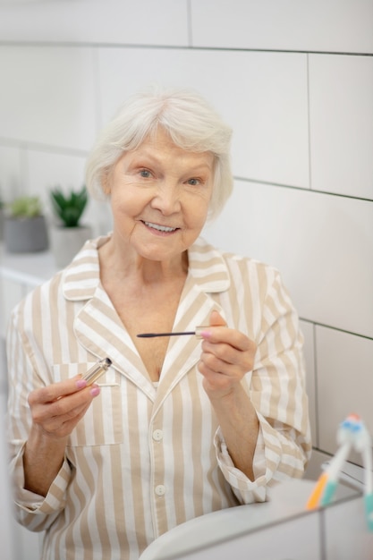 Maquillaje. Mujer de pelo gris en pijama con rímel en las manos