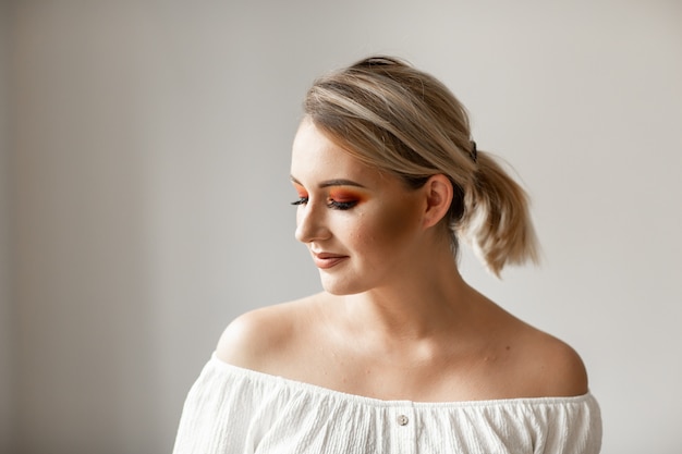 Maquillaje de prueba en un salón de belleza. Joven rubia con cola de caballo y maquillaje brillante sobre fondo liso. Espacio para texto