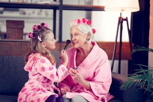 Maquillarse juntos. Niña y abuela vistiendo rulos y batas de baño rosa haciendo maquillaje juntos