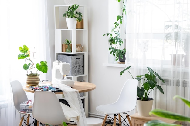 Máquina de costura em uma mesa redonda no interior branco da casa perto da janela com uma cortina transparente e uma planta caseira em uma panela, um negócio doméstico de passatempo interior moderno