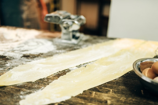 Máquina de macarrão, massa e ovos na mesa da cozinha de madeira polvilhada com farinha, ninguém. Cozinha tradicional italiana