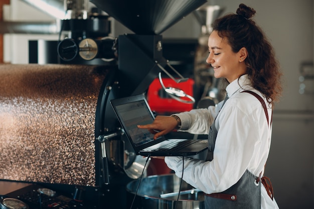 Máquina torradeira de café e mulher barista com computador portátil no processo de torrefação de café.