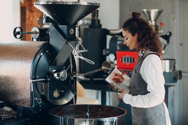 Máquina tostadora de café y mujer barista con lápiz de escritura de tableta en el proceso de tostado de café