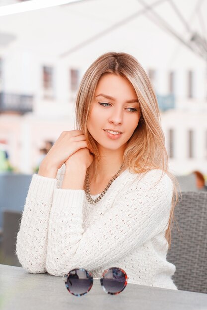 Maravilhosa bela jovem com um suéter de malha vintage de malha com olhos azuis está sentado e sonhando em um café ao ar livre de verão em um dia ensolarado. Menina encantadora gosta do resto.