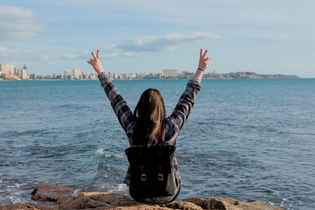 Maravilhosa e alegre jovem viajante levanta os braços feliz olha para o mar ao ar livre em um dia ensolarado