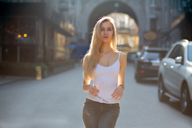 Maravilhosa mulher loira com cabelos longos, vestindo jeans e camiseta, andando na cidade sob os raios de sol