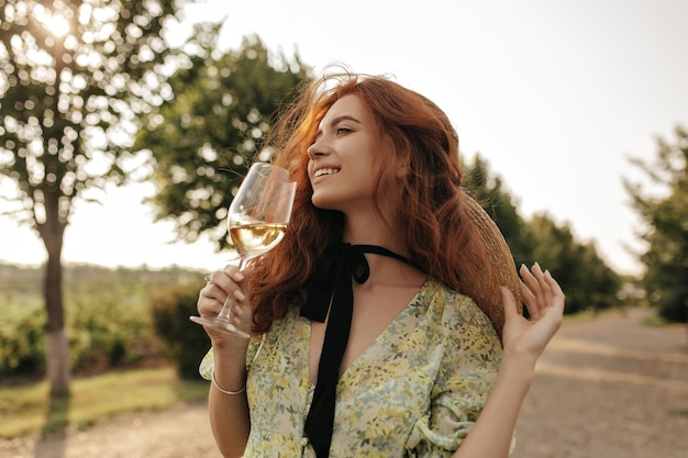 Maravilhosa mulher ruiva de bom humor com chapéu de palha e vestido verde moderno sorrindo e segurando copo com vinho ao ar livre