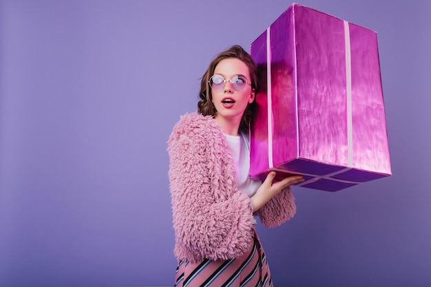 Maravillosa cumpleañera en abrigo de piel sosteniendo un gran regalo Toma interior de una encantadora dama europea posando con regalo en el estudio