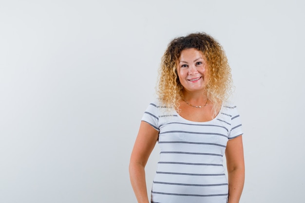 Maravillosa dama en camiseta a rayas sonriendo mientras está de pie y mirando alegre, vista frontal.