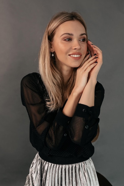 Maravillosa linda chica europea con cabello rubio usando blusa negra y falda plateada tocando suavemente su rostro y mirando hacia otro lado con una maravillosa sonrisa sobre fondo gris