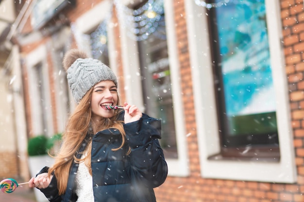 Maravillosa mujer joven disfrutando de las vacaciones de invierno y mordiendo piruletas de colores en la calle. Espacio vacio