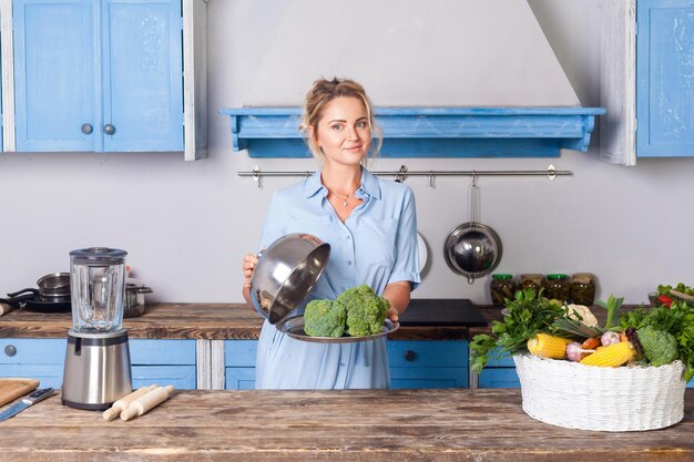 Maravillosa mujer joven sosteniendo brócoli en la bandeja debajo de la campana y mirando a la cámara hogar mostrando verduras cocidas comida vegetariana saludable nutrición vegana y vitaminas cocina moderna interior