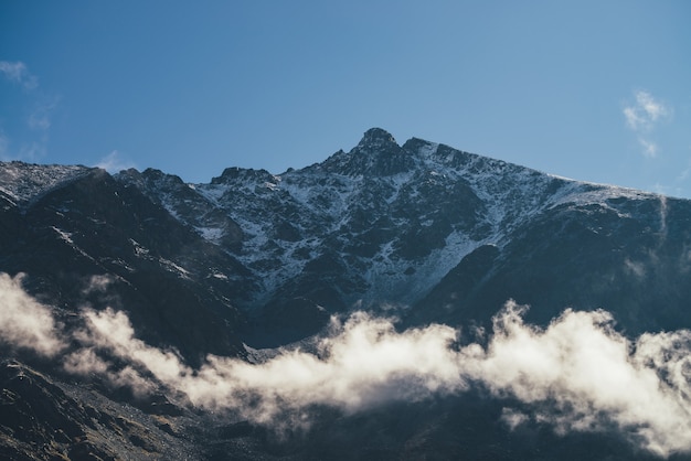 Maravillosa vista de la pared de la montaña rocosa con nieve por encima de las nubes y pico. Pintoresco paisaje de montaña con pico puntiagudo y nubes bajas sobre rocas. Paisaje alpino mínimo con pináculo rocoso afilado.