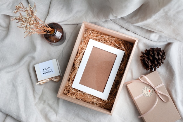 Un marco blanco colocado en una caja de regalo marrón rodeada por una caja de pequeños anillos de diamantes atados con piñas de color crema, flores secas en una botella de vidrio marrón.