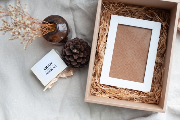 Un marco blanco colocado en una caja de regalo marrón rodeada por una caja de pequeños anillos de diamantes atados con piñas de color crema, flores secas en una botella de vidrio marrón.