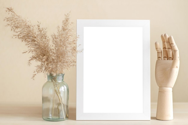 Un marco blanco vacío se alza sobre una mesa de madera beige con hierbas secas en un jarrón y una mano de madera.