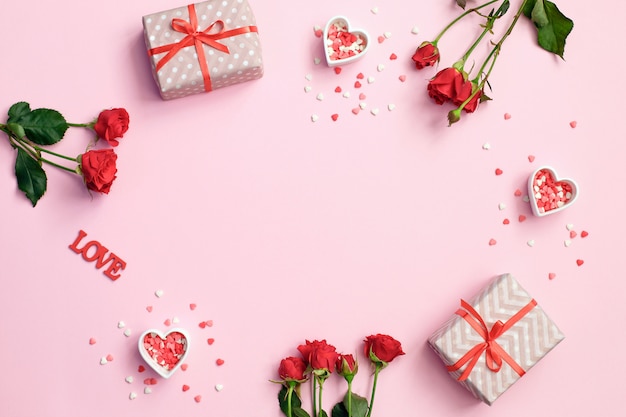 Marco del día de San Valentín hecho de flores color de rosa, corazones de regalos sobre fondo rosa