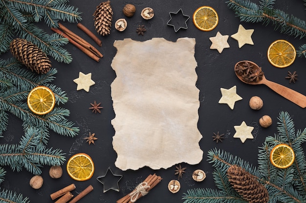 Marco festivo con papel vintage y adornos navideños. Abeto, naranja, canela, galletas de jengibre y tarjeta en blanco sobre fondo negro, espacio de copia