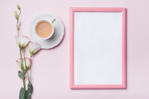 Foto marco de fotos en blanco con borde rosa; taza de té y flores frescas de eustoma contra el fondo coloreado