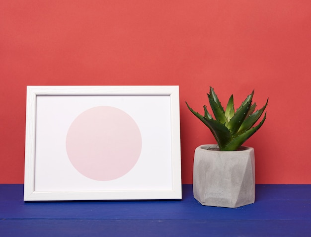 Marco de fotos blanco y maceta de cerámica con una planta en crecimiento sobre una mesa de madera azul