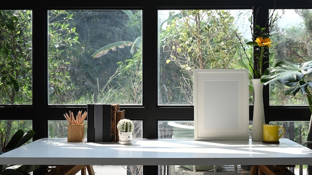 Marco de fotos, libros de tazas de café y maceta en una mesa blanca cerca de una ventana de vidrio en la oficina del hogar