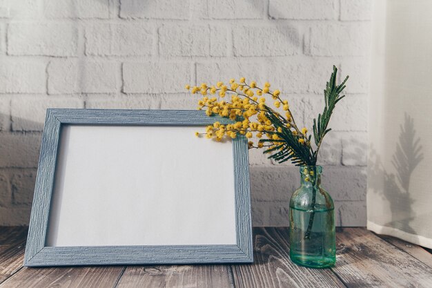 Marco de fotos y una pequeña botella con flores de mimosa en la superficie de una pared de ladrillos