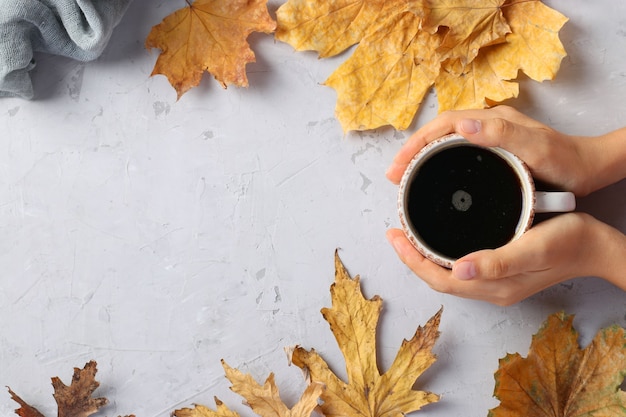 Marco con hojas de otoño y una taza de café en manos femeninas sobre un fondo gris, vista superior, plano, espacio de copia