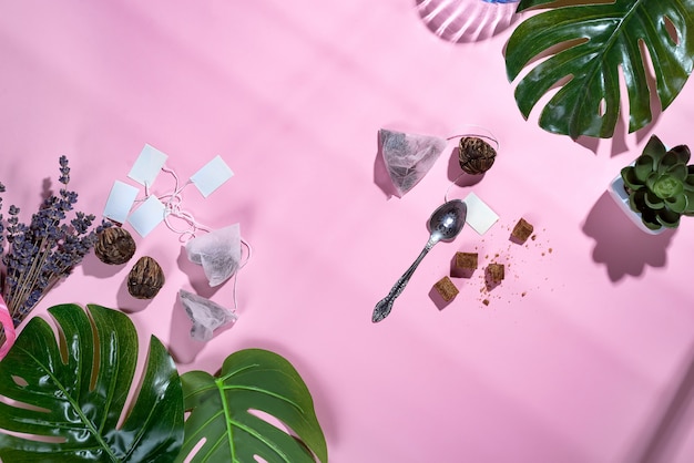 Marco con hojas tropicales verdes y taza de té, bolsa de té y azúcar sobre fondo rosa pastel