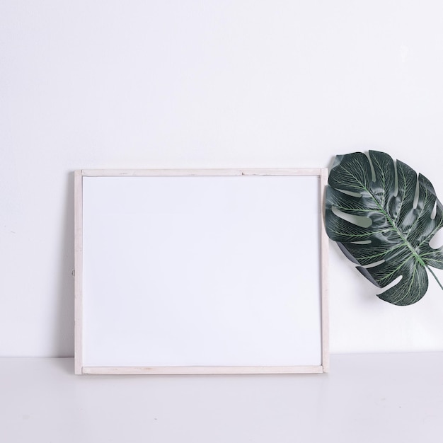 Marco horizontal de madera en blanco y hoja artificial en la mesa con fondo de pared blanca. Tema de la maqueta