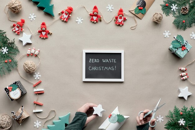 Marco de Navidad cero desperdicio con espacio de copia. Vista plana endecha, superior en papel artesanal marrón. Baratijas textiles, árboles de hoja perenne, caja de regalo de papel en la mano. Eco amigable Navidad, "Zero Waste Christmas" en la pizarra.