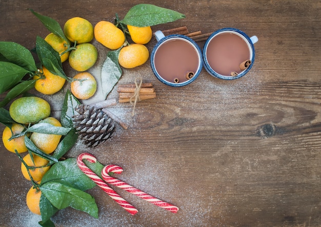 Foto marco de navidad o año nuevo. mandarinas frescas con hojas, palitos de canela, piña, chocolate caliente en tazas y bastones de caramelo sobre fondo de madera rústica, vista superior