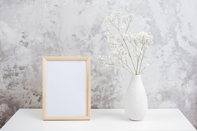 Marco vacío blanco vertical de madera y ramo de flores pequeñas blancas gypsophila en jarrón sobre mesa en muro de hormigón gris