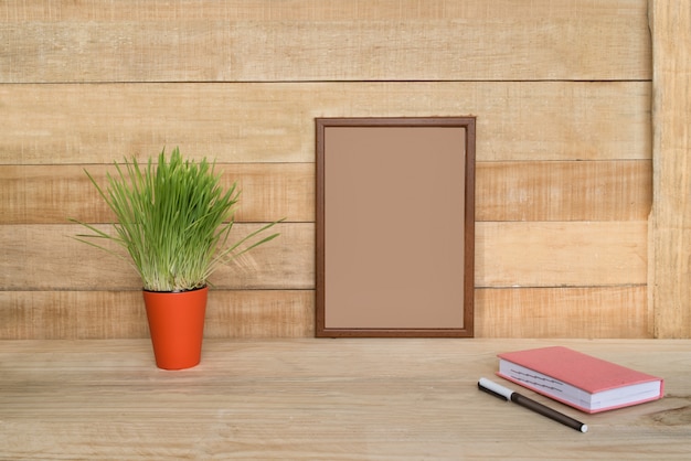 Marco vacío en una pared de madera. Cuaderno y bolígrafo, planta de interior verde sobre la mesa. Espacio de trabajo en casa