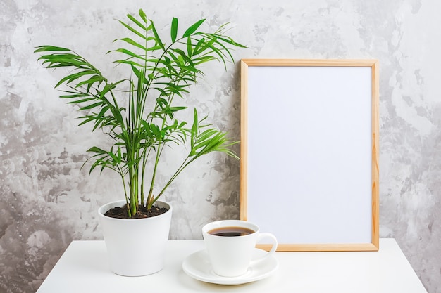 Marco vertical de madera con tarjeta blanca en blanco, taza de café y flor de palma exótica verde en maceta en la mesa sobre muro de hormigón gris