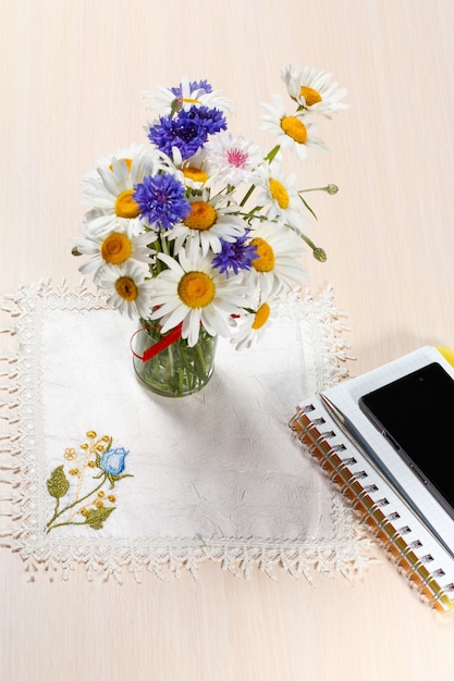 Margarida camomila e flores em um vaso em uma mesa de madeira com um guardanapo, caderno, celular e uma caneta