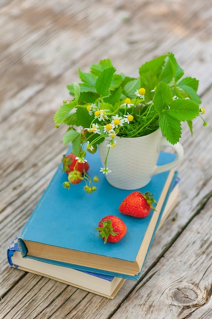 Margaritas silvestres y fresas en una taza blanca en libros sobre una vieja mesa de madera rústica