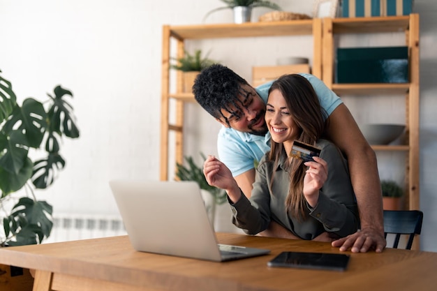 Marido abraçando sua esposa enquanto ela usa laptop e cartão de crédito em casa
