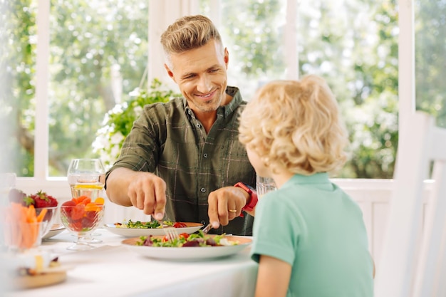 Marido bonito sorrindo enquanto corta salada para seu filho encaracolado